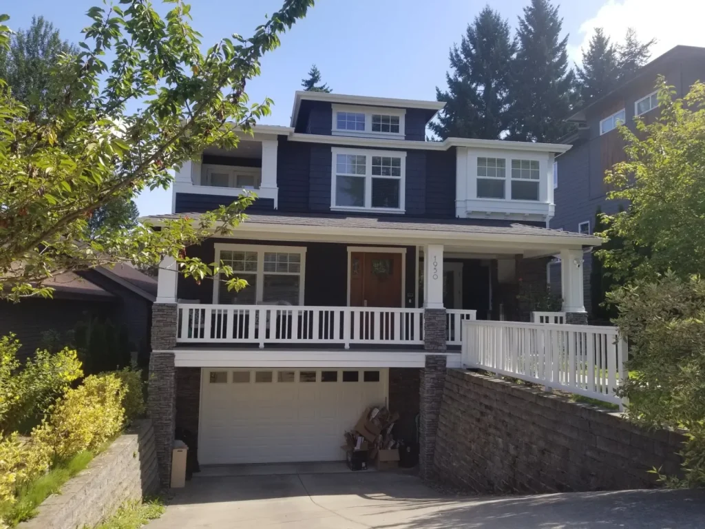 Two-story house with dark siding, white trim, a front porch, and a double garage on a sloped driveway.