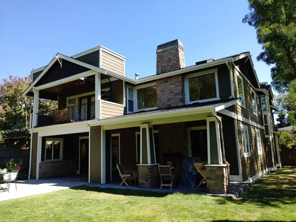 Two-story modern house with stone chimney, covered patio, balcony, and green lawn under a clear blue sky.