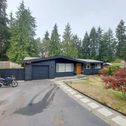 Single-story dark blue house with attached garage, driveway, motorcycle, and surrounding trees on an overcast day.