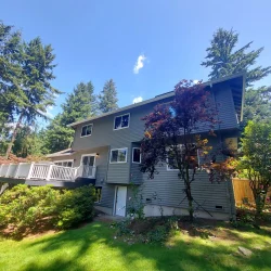 Two-story gray house with deck, surrounded by green grass, trees, and shrubs on a sunny day.