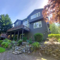 Two-story dark-colored house with large windows, a wooden balcony, and landscaped garden with stone retaining wall.