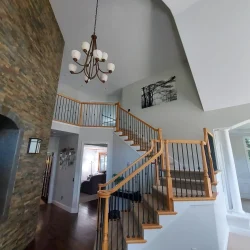 A wooden staircase with black railings leads to an upstairs landing in a modern home with high ceilings and a chandelier.