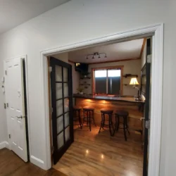 A small home bar with three bar stools, wood flooring, and warm lighting, viewed through open double doors.
