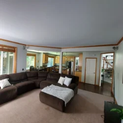 Spacious living room with brown sectional sofa, large windows, and a view of the kitchen and staircase in the background.