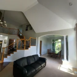Modern living room with a black sofa, open staircase, and dining area with tall windows and natural light.