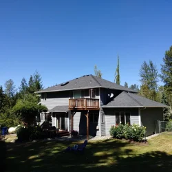 Two-story gray house with a small balcony, surrounded by trees and a grassy yard under a clear blue sky.