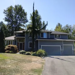 Two-story gray house with a red front door, surrounded by trees and shrubs, next to a driveway on a sunny day.