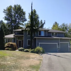 Two-story gray house with a red front door, surrounded by trees and shrubs, next to a driveway on a sunny day.