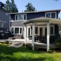 Two-story house with dark siding, a raised wooden deck, outdoor seating, and a covered patio in a grassy backyard.