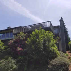 Two-story house with large balcony, surrounded by green bushes and trees under a clear sky.