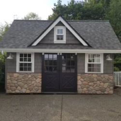 A small gray and stone outbuilding with double doors, two windows, and a gabled roof, set on a paved driveway.
