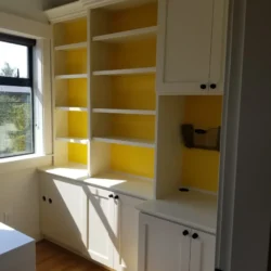 White built-in cabinets and shelves with a yellow back panel, next to a window with sunlight streaming in.