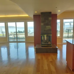 Bright living room with hardwood floors, modern fireplace, large windows, and a view of water and industrial structures outside.