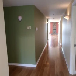 A hallway with light wood floors, green and white walls, and wall-mounted lights leads to a room with a fireplace.