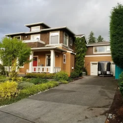 Two-story house with brown siding, attached garage, front porch, and landscaped yard under a cloudy sky.