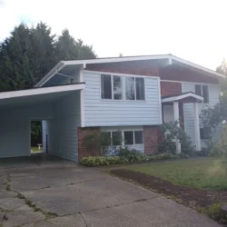 Two-story house with light blue siding, brown accents, large windows, a carport, and a concrete driveway.