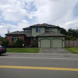 Two-story suburban house with green siding, double garage, brick accent, and a car parked on the street in front.