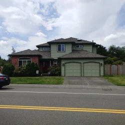 Two-story suburban house with green siding, double garage, brick accent, and a car parked on the street in front.