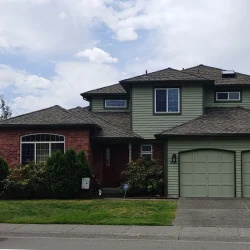 Two-story suburban house with green siding, brick accent, double garage, and a blue car parked on the street.