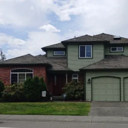 Two-story suburban house with green siding, brick accent, double garage, and a blue car parked on the street.