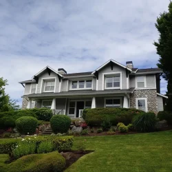 Two-story white house with covered porch, stone accents, and neatly landscaped front yard under a partly cloudy sky.