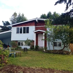 Two-story house with white siding and red accents, front yard with lawn chairs and trees, and a flower bush.