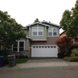 Two-story suburban house with a double garage, trees, and trash bins on the driveway, photographed from the street.