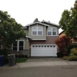Two-story suburban house with a double garage, trees, and trash bins on the driveway, photographed from the street.