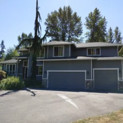 Two-story gray house with three-car garage, surrounded by trees and a paved driveway under clear skies.