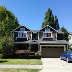 Two-story suburban house with brown siding, a double garage, and a blue car parked in the driveway on a sunny day.