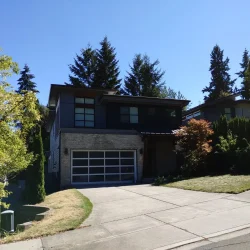 Modern two-story house with large windows, a double garage, and a sloped driveway surrounded by trees and grass.