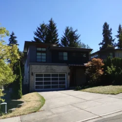 Modern two-story house with large windows, a double garage, and a sloped driveway surrounded by trees and grass.