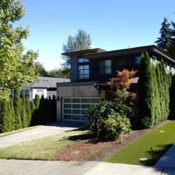 Modern two-story house with large windows, a glass garage door, and landscaped front yard with trees and shrubs.